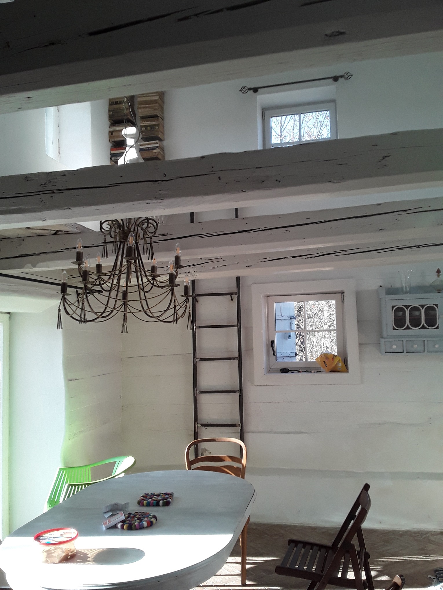 Jasne wnętrze z białymi ścianami i drewnianymi belkami stropowymi, oświetlone żyrandolem, z drabiną opartą o ścianę i widocznymi elementami dekoracyjnymi.