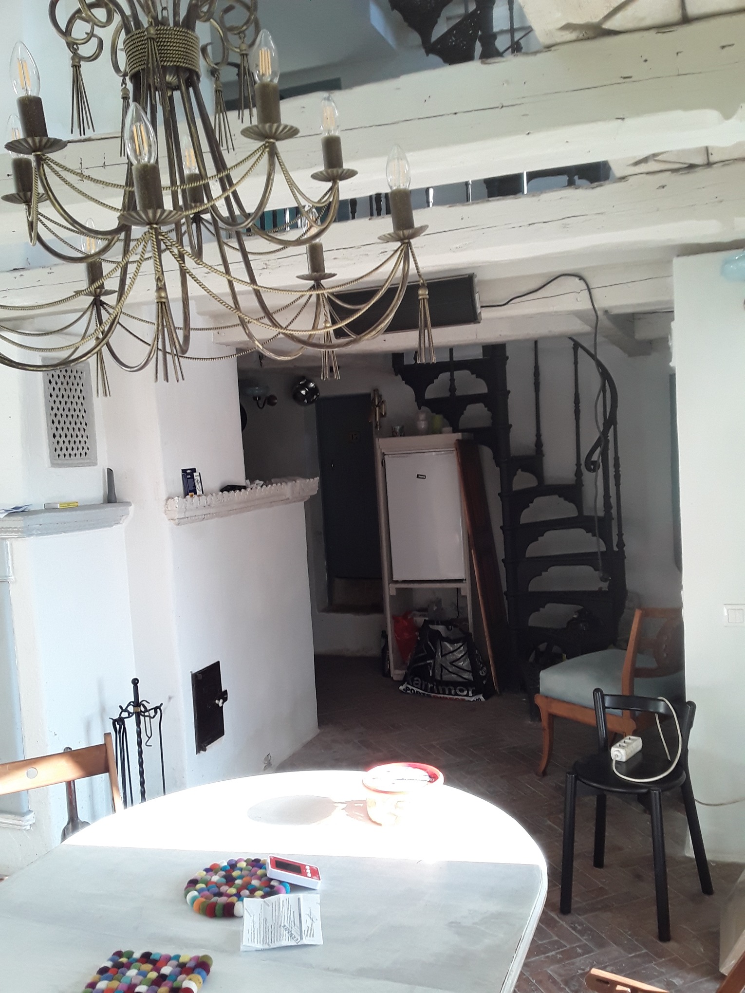 Jasne wnętrze z białymi ścianami, ozdobnym żyrandolem i spiralnymi schodami. Widoczny piec kaflowy, stół z dekoracjami i krzesła. Charakterystyczne białe belki stropowe.