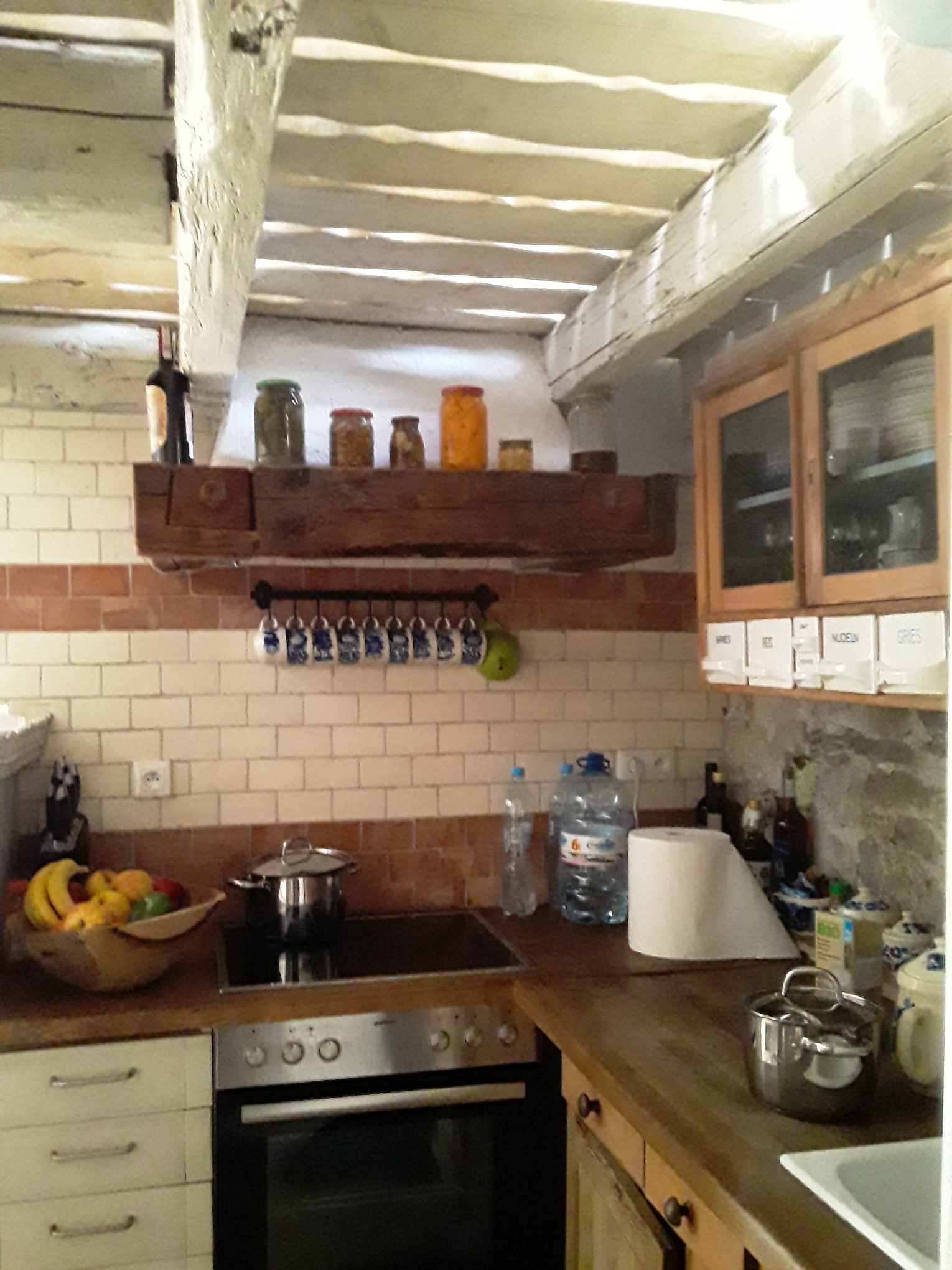 Rustykalna kuchnia z drewnianym sufitem, ceglaną glazurą nad blatem, drewnianymi szafkami i półkami, oraz różnorodnymi akcesoriami kuchennymi.