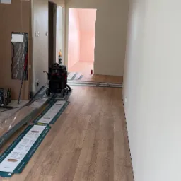 Jasne panele podłogowe ułożone w korytarzu, widoczne narzędzia pracy i odkurzacz przemysłowy. Świeżo wykończone ściany w neutralnym odcieniu.