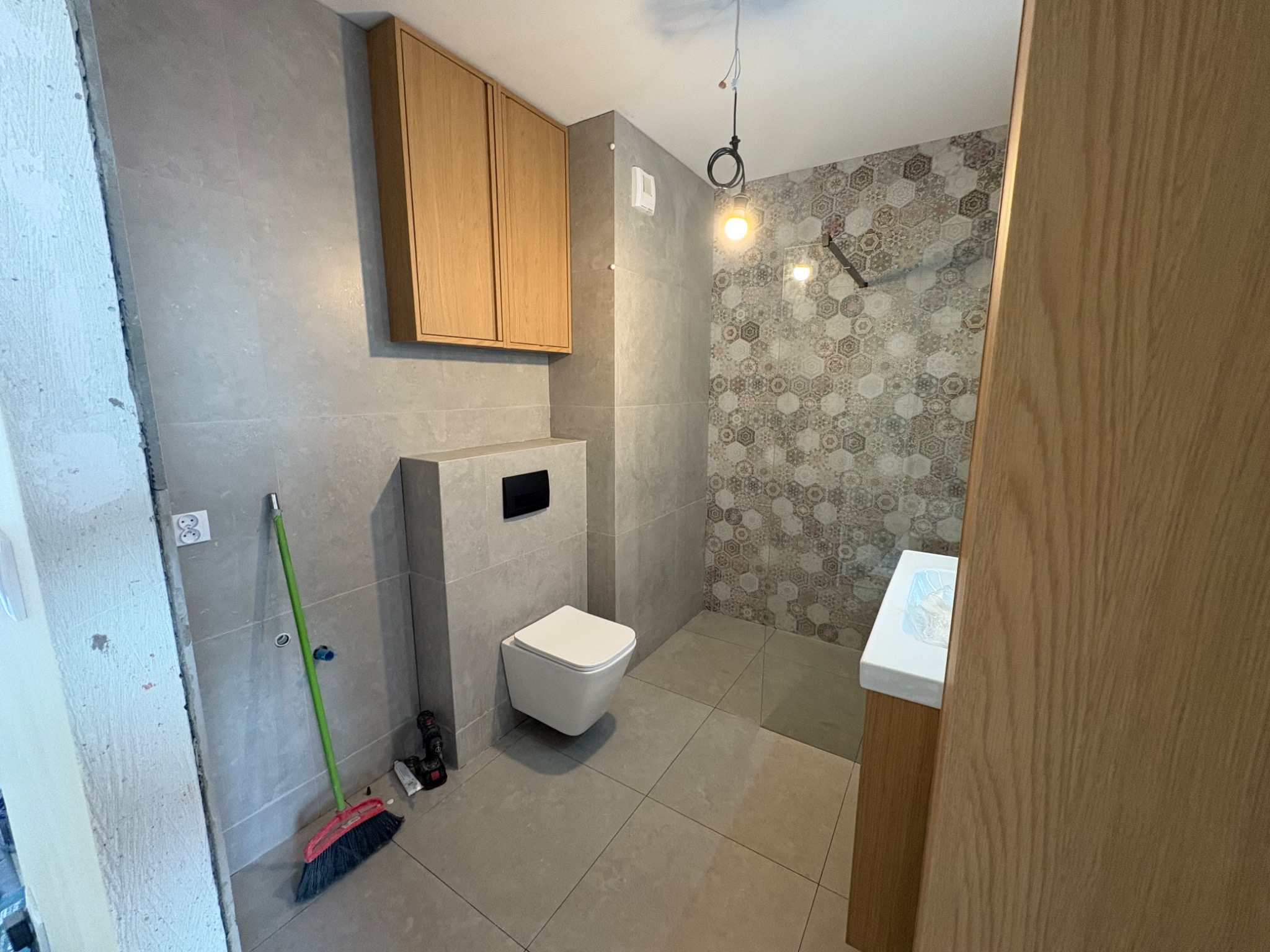 Wykończona łazienka z szarą płytką i heksagonalnym wzorem pod prysznicem. Wisząca toaleta i drewniane szafki nad nią. Widoczna miotła i narzędzia na podłodze.