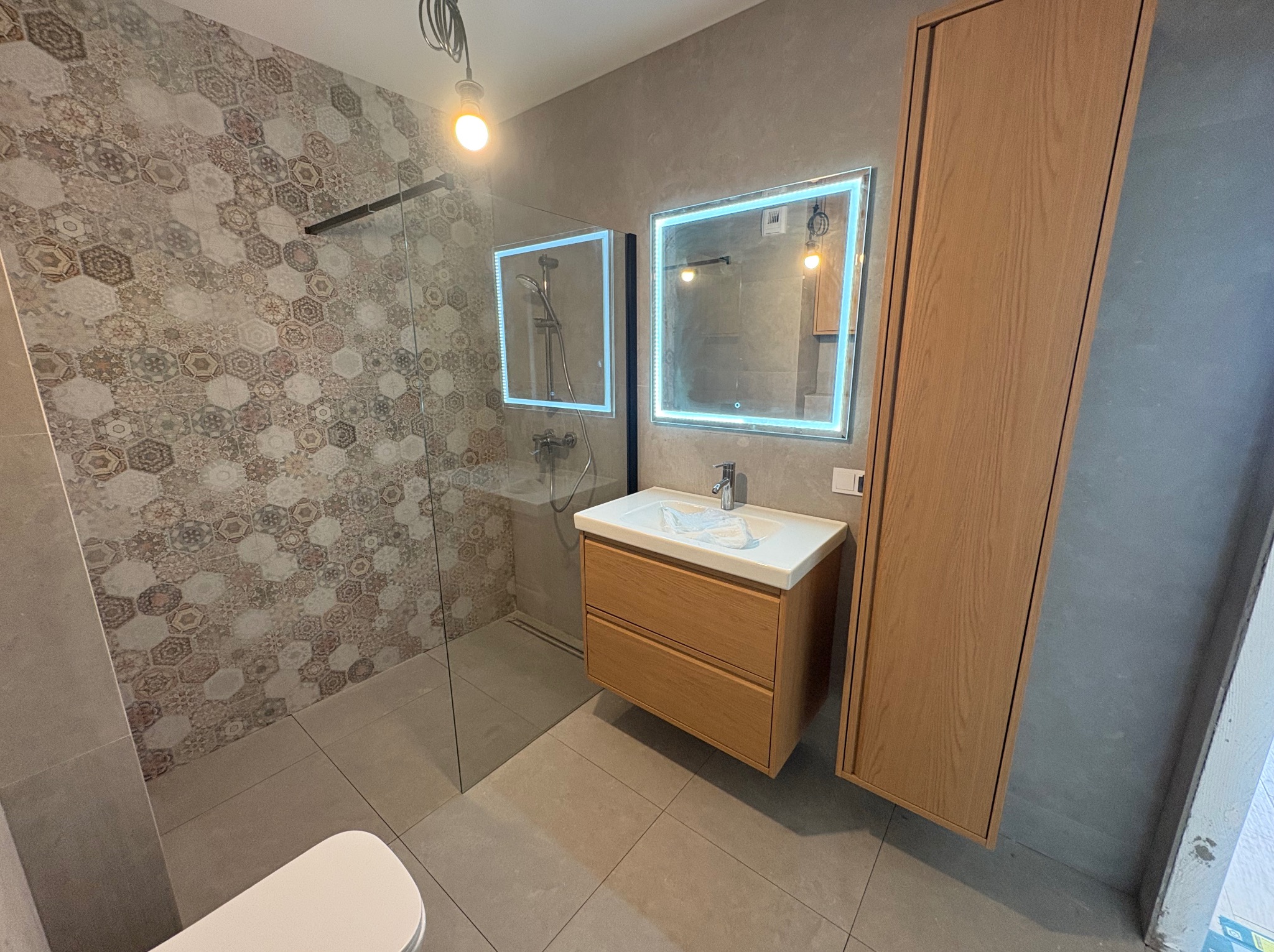 Nowoczesna łazienka z heksagonalną mozaiką pod prysznicem, drewnianą szafką z umywalką i lustrem z podświetleniem LED. Minimalistyczny design.