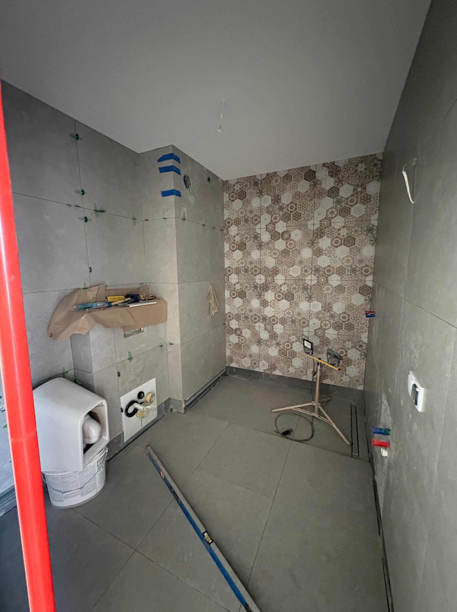 Łazienka w trakcie prac: ułożone płytki w szarościach i wzorzyste heksagony na ścianie, narzędzia na obudowie, poziomica na podłodze, lampa budowlana.
