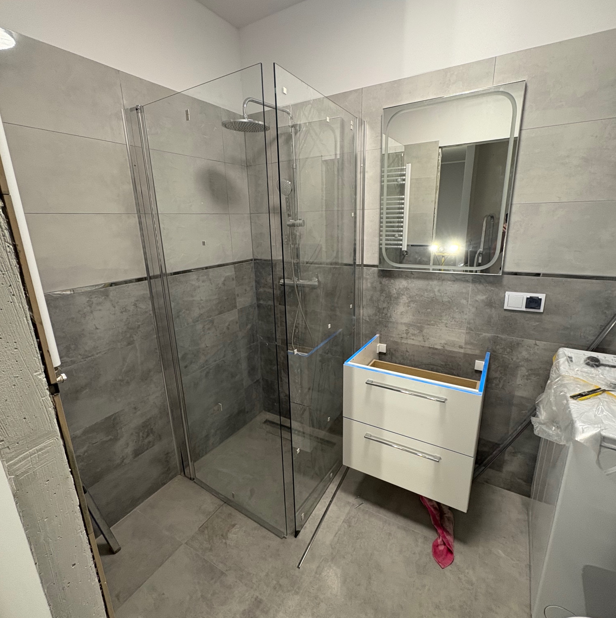 Szara łazienka w trakcie wykańczania: kabina prysznicowa ze szklanymi ścianami, szafka pod umywalkę bez blatu. Widoczne płytki i lustro z podświetleniem LED.