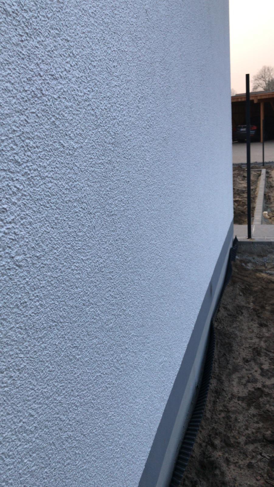 Fragment ściany zewnętrznej budynku z szarą, ziarnistą fakturą tynku, widoczny cokół i fragment otoczenia z czarną kratką drenażową przy fundamencie.