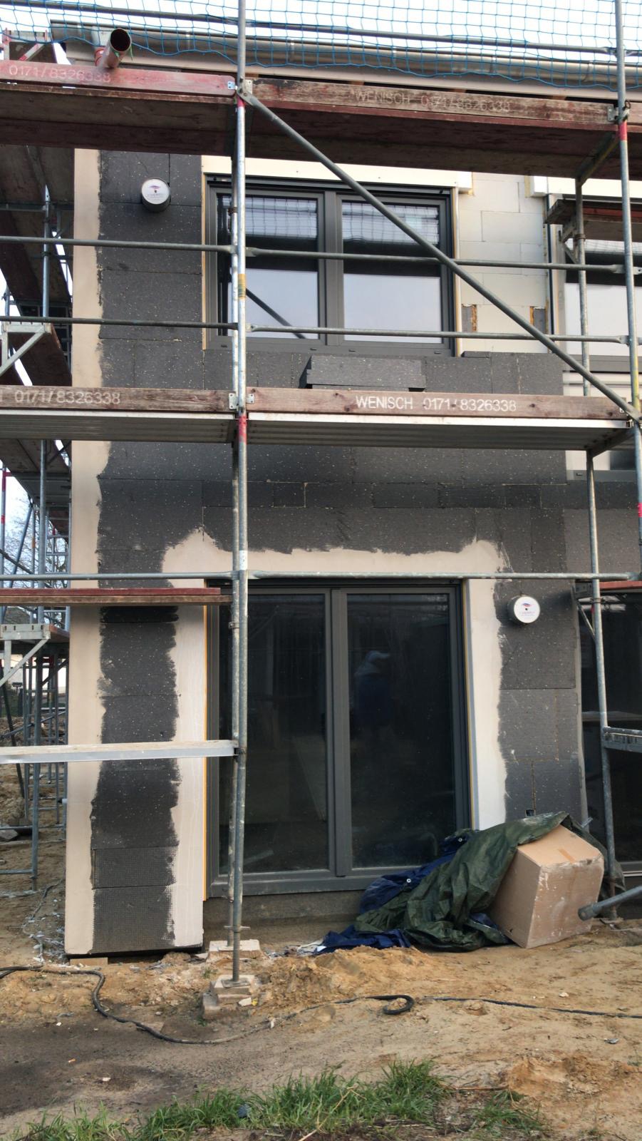 Budynek w trakcie ocieplania, widoczne rusztowania, szare bloczki styropianowe, częściowo nałożona warstwa szpachli, okno w szarej ramie, ziemia i trawa u podstawy budynku.