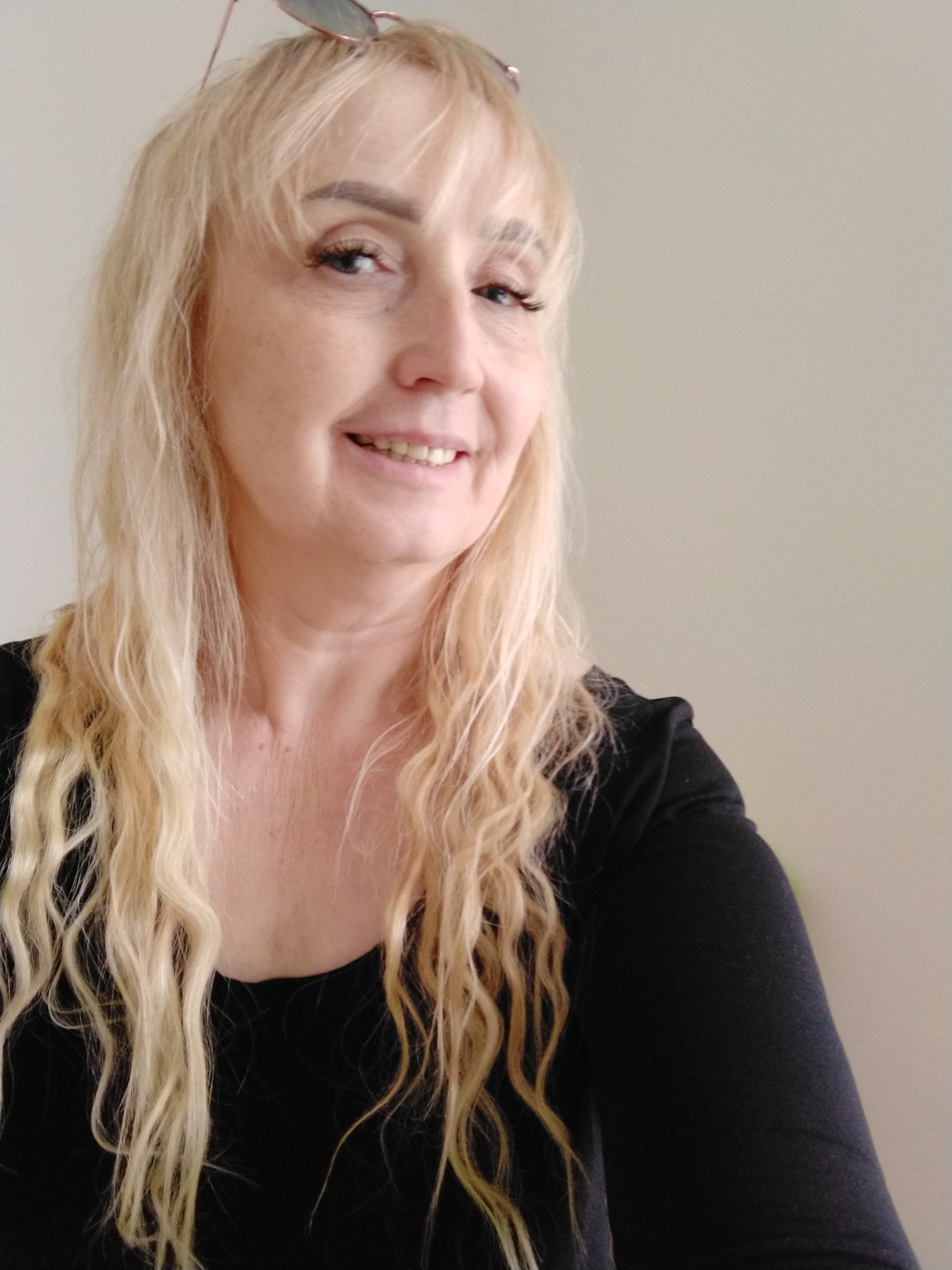 Portret uśmiechniętej kobiety z blond, falowanymi włosami i okularami na głowie, ubrana w czarną bluzkę na neutralnym tle.