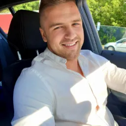 Uśmiechnięty mężczyzna w białej koszuli siedzi w samochodzie, z zewnątrz widoczne drzewa i fragment innego pojazdu. Jasne, naturalne światło.