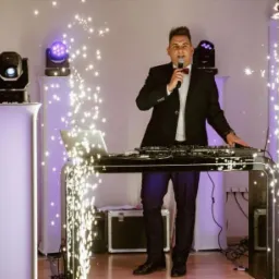 Mężczyzna w garniturze z mikrofonem, stojący za konsoletą DJ-ską otoczoną iskrami, obok kolumny oświetleniowe z reflektorami.