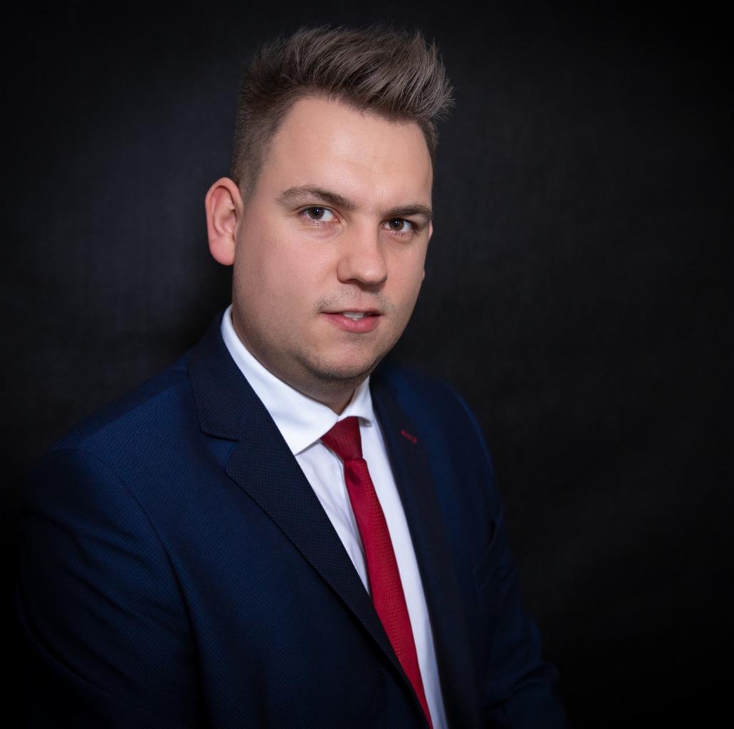 Portret mężczyzny w niebieskiej marynarce i czerwonym krawacie na czarnym tle, z delikatnym uśmiechem.