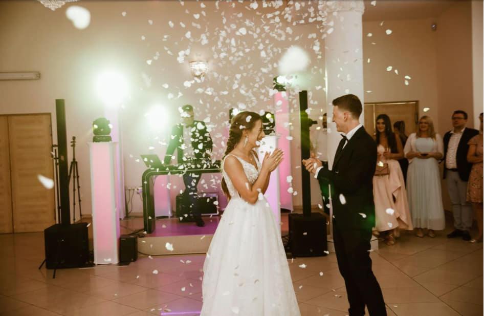 Para młoda w trakcie pierwszego tańca, obsypywana płatkami kwiatów, w tle DJ na podświetlonej scenie i goście.