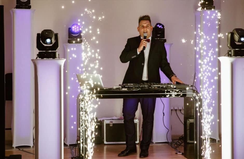 Mężczyzna w garniturze z mikrofonem, stojący za konsoletą DJ-ską otoczoną iskrami, obok kolumny oświetleniowe z reflektorami.
