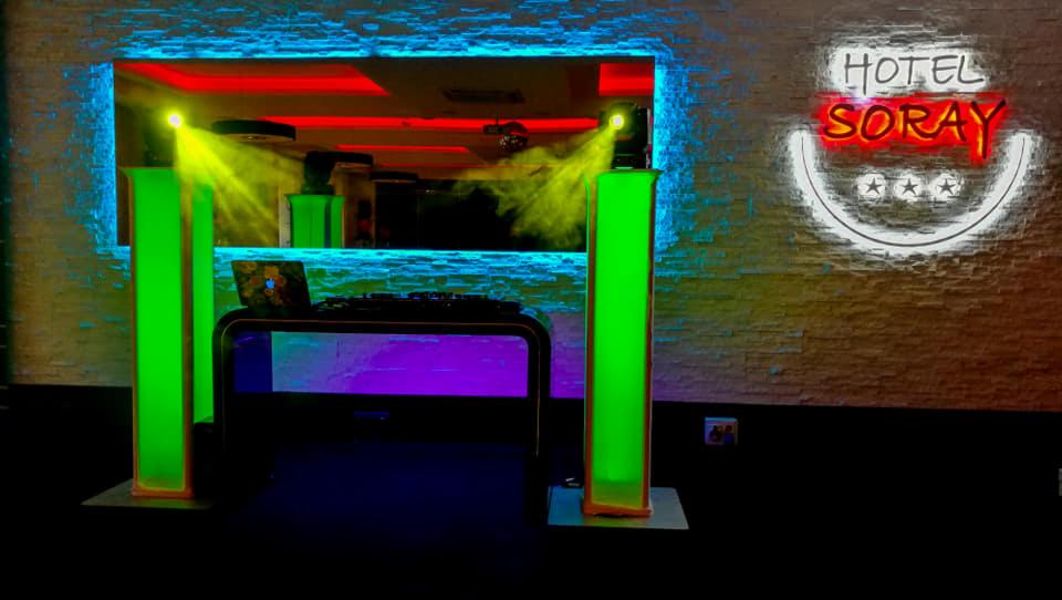 Stanowisko DJ-a z laptopem na stole, otoczone zielonymi kolumnami oświetleniowymi i reflektorami, całość w neonowym oświetleniu wnętrza z logo 'Hotel Soray' na ścianie.