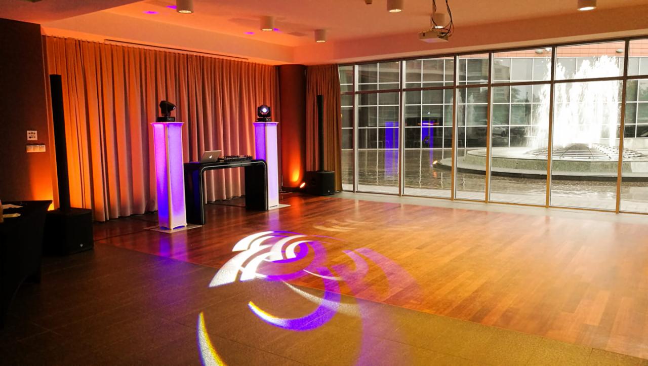 Sala z parkietem przygotowana na imprezę: kolumny głośnikowe, konsoleta DJ-ska, oświetlenie punktowe skierowane na parkiet, za oknami fontanna.