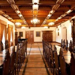 Długa sala konferencyjna z drewnianym sufitem kasetonowym, parkietem i dwoma długimi stołami otoczonymi rzeźbionymi krzesłami. W tle widoczne zabytkowe meble i okna z zasłonami.