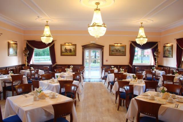 Elegancka sala bankietowa w jasnych barwach z drewnianymi krzesłami, nakrytymi białymi obrusami stołami, kryształowymi żyrandolami i dekoracyjnymi obrazami na ścianach.