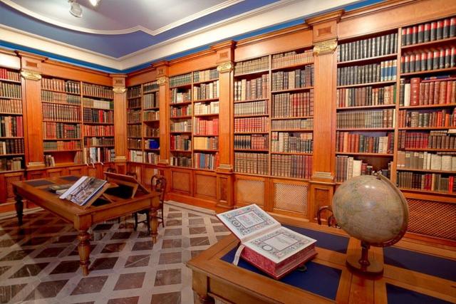 Bogato wyposażona biblioteka z drewnianymi regałami wypełnionymi książkami, drewnianym biurkiem z otwartą księgą i globusem, oraz marmurową podłogą.