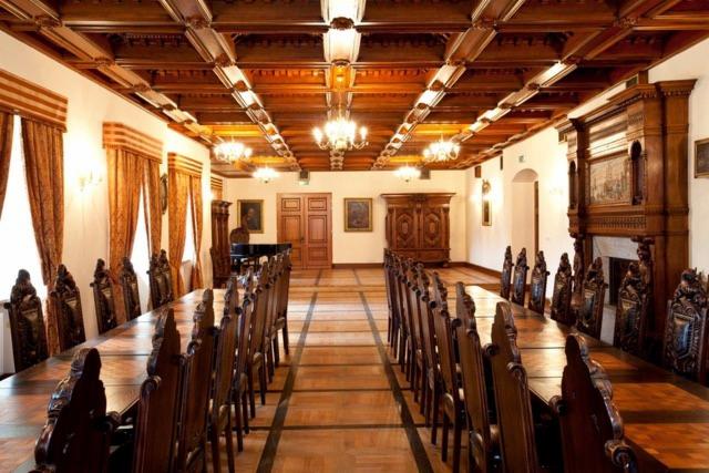 Długa sala konferencyjna z drewnianym sufitem kasetonowym, parkietem i dwoma długimi stołami otoczonymi rzeźbionymi krzesłami. W tle widoczne zabytkowe meble i okna z zasłonami.