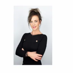 Portret uśmiechniętej kobiety z upiętymi włosami, w czarnej sukience z ozdobnymi złotymi guzikami i zegarkiem na ręce, na jasnym tle.