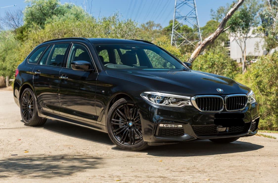 Czarny samochód kombi marki BMW z czarnymi felgami, zaparkowany na betonowej nawierzchni na tle zieleni i słupa wysokiego napięcia.