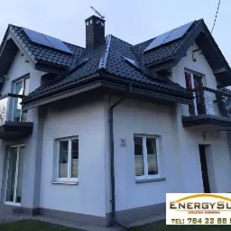 Dom jednorodzinny z panelami słonecznymi na dachu, widoczne balkony z przeszklonymi balustradami, elewacja w odcieniach szarości, logo firmy w prawym dolnym rogu.