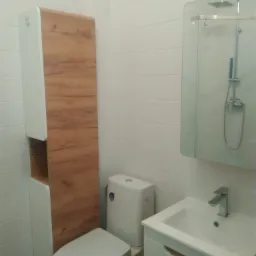 Biała łazienka z toaletą, umywalką z szafką i wysoką szafką z drewnianym frontem, widoczne białe płytki na ścianach i lustro z kabiną prysznicową.