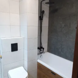 Nowoczesna łazienka z białymi płytkami na ścianach, podwieszaną toaletą z czarnym przyciskiem, czarną armaturą prysznicową i wanną obudowaną drewnopodobną płytką.