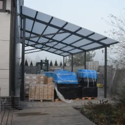 Zadaszenie z poliwęglanu na metalowej konstrukcji wspartej na słupach, pod którym składowane są palety z materiałami budowlanymi, w tle widoczny wózek widłowy oraz ogrodzenie.