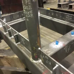 Szczegółowy widok spawanej konstrukcji stalowej z widocznymi numerami i oznaczeniami na elementach, niebieskie zaślepki zabezpieczające otwory, ślady po spawaniu.