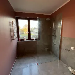 Nowoczesna łazienka z kabiną prysznicową typu walk-in, szare płytki podłogowe i częściowo ścienne, ściany pomalowane na kolor brudny róż, grzejnik drabinkowy przy oknie.