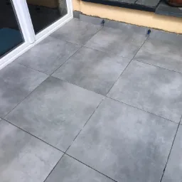 Szare, kwadratowe płytki na balkonie, widoczne niebieskie elementy poziomujące, przesuwne drzwi balkonowe w tle.