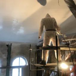 Pracownik w kombinezonie ochronnym, stojący na rusztowaniu, aplikuje gładź natryskową na suficie pomieszczenia z oknami w kształcie łuku i niezabezpieczonymi elementami konstrukcyjnymi.