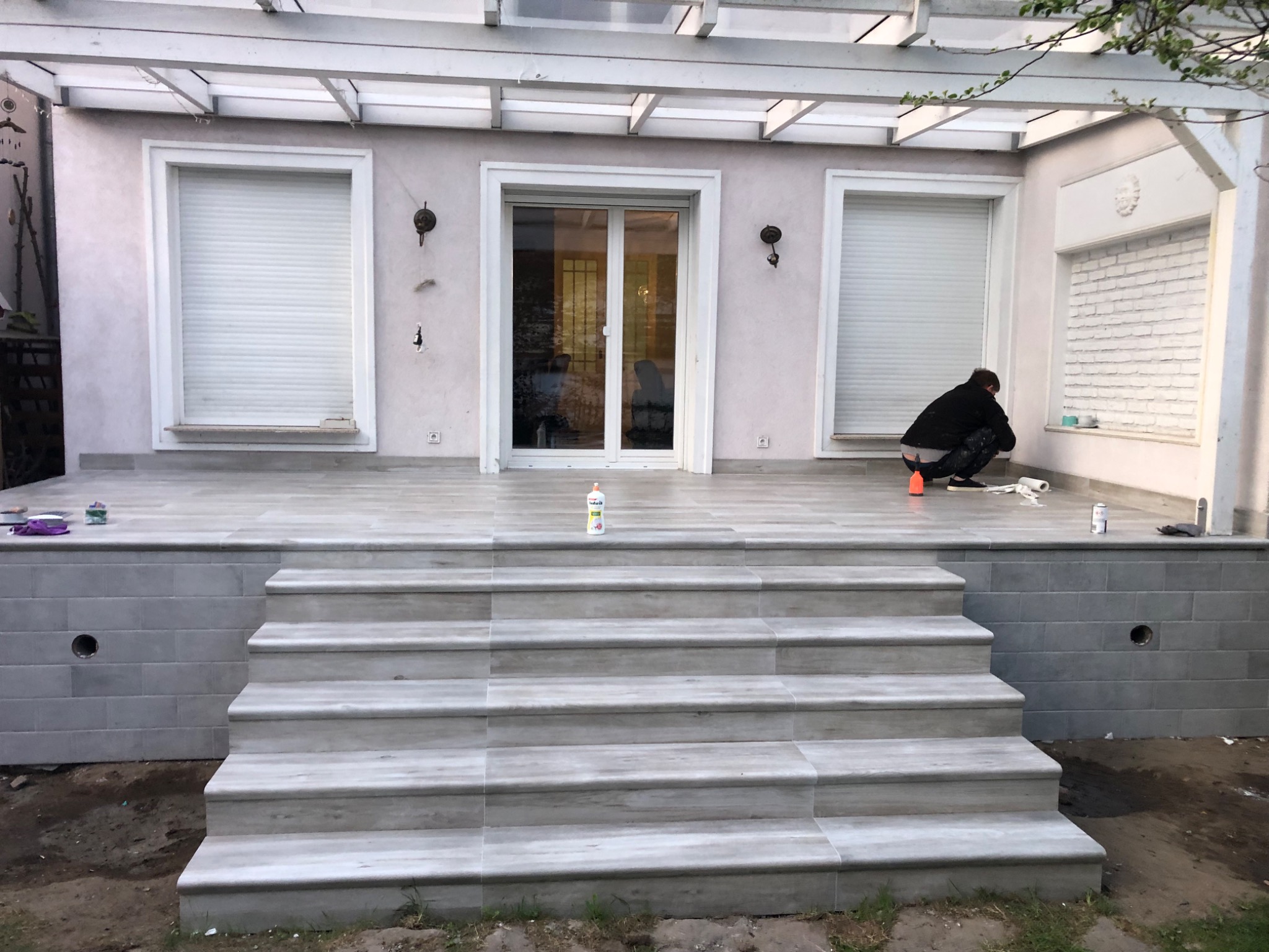 Wyłożony płytkami taras z schodami, widoczna osoba pracująca przy ścianie budynku, biała pergola nad tarasem.