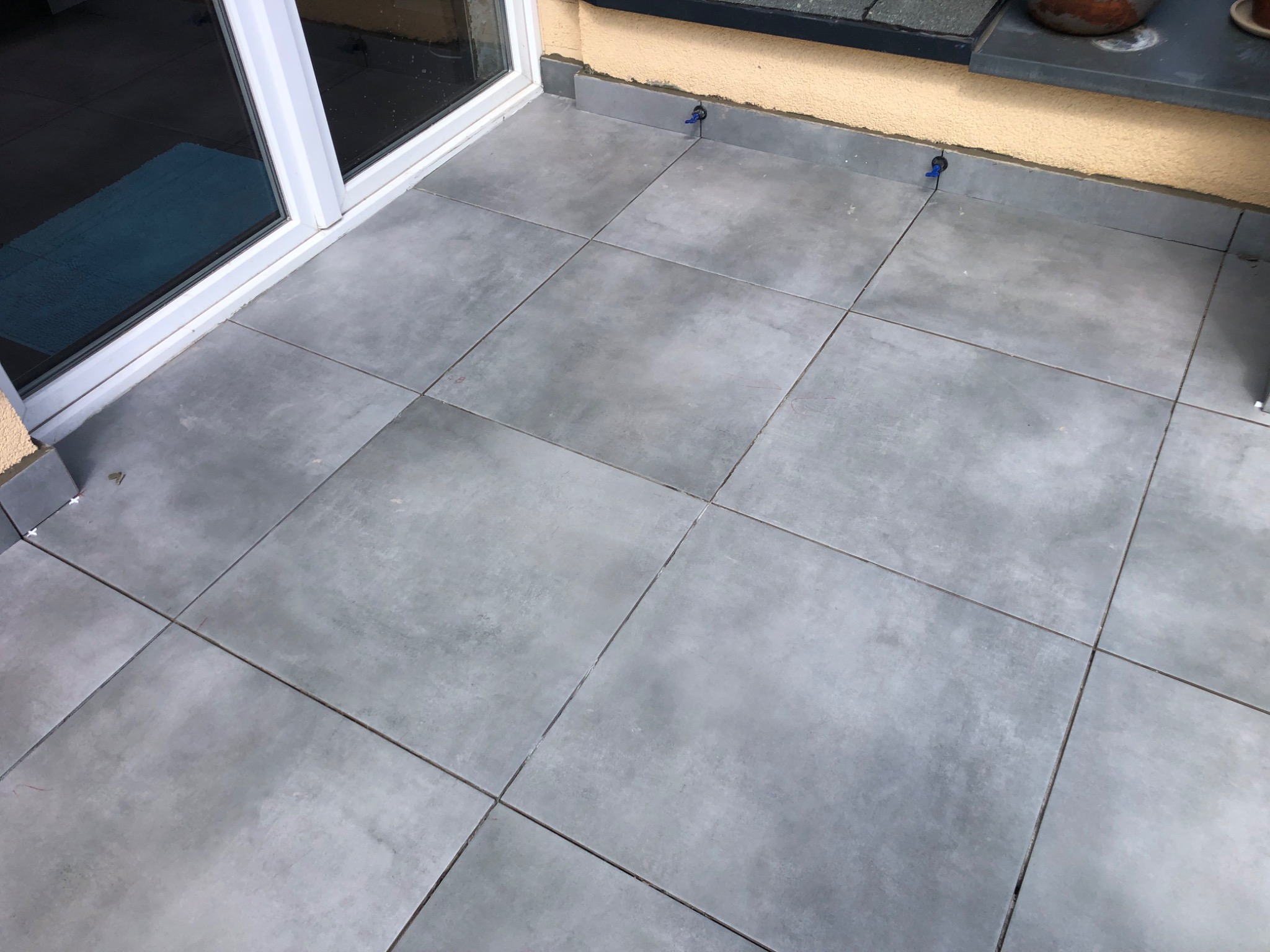 Szare, kwadratowe płytki na balkonie, widoczne niebieskie elementy poziomujące, przesuwne drzwi balkonowe w tle.