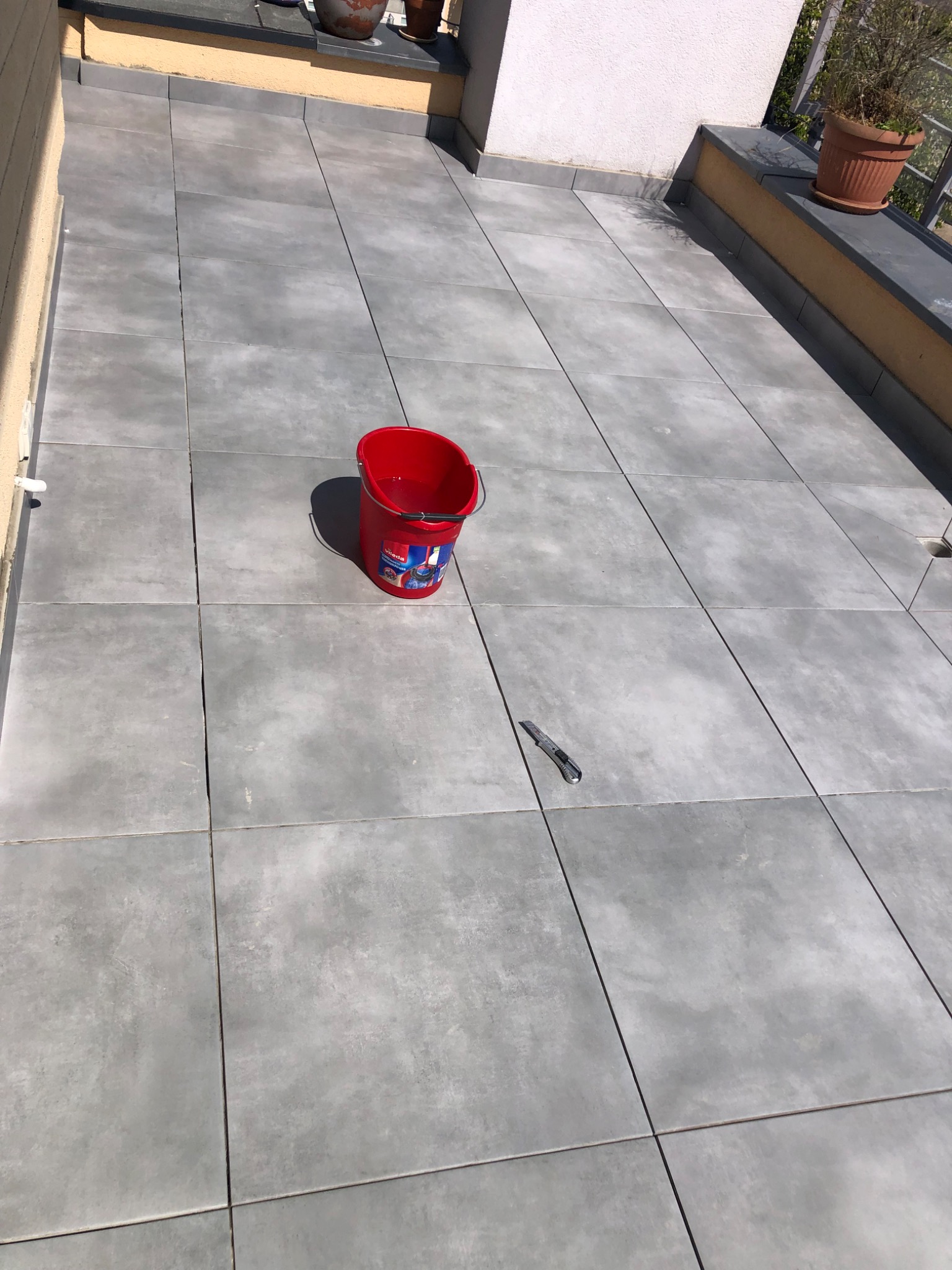 Szare płytki podłogowe na balkonie, czerwone wiadro Vileda z wodą i nóż do tapet leżący na płytkach.