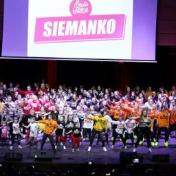 Duża grupa dzieci i młodzieży w kolorowych strojach tanecznych na scenie podczas występu, w tle widoczny ekran z logo 'Studio Tańca Siemanko'.