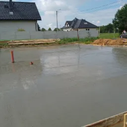 Świeżo wylana, mokra płyta betonowa pod budynek, otoczona drewnianym szalunkiem. W tle widoczny dom z szarą elewacją i ciemnym dachem, oraz zaparkowany samochód osobowy.