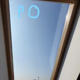 Okno dachowe z drewnianą ramą, widok na błękitne niebo i odległe drzewa, na szybie widoczne niebieskie ślady po czyszczeniu.