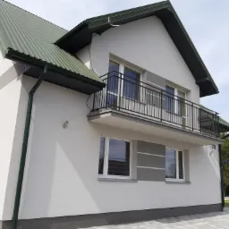 Dwukondygnacyjny dom z balkonem, jasną elewacją z szarymi akcentami, zielonym dachem z blachy trapezowej i ciemnozielonymi rynnami, widok z zewnątrz na kostkę brukową.