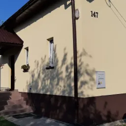 Elewacja domu jednorodzinnego z numerem 147, ocieplona i pomalowana na beżowo z brązowym cokołem, widoczny zadaszony ganek z kwiatami i małe okna.