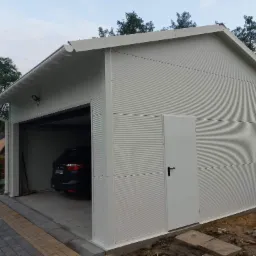 Biały garaż z płyt warstwowych z otwartą bramą, wewnątrz zaparkowany ciemny samoch&oacute;d, widoczne rynny i drzwi wejściowe z boku.