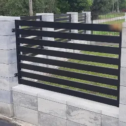 Nowoczesne, poziome, czarne przęsło ogrodzeniowe zamontowane pomiędzy słupkami z szarej kostki betonowej. Widoczna trawa w tle.