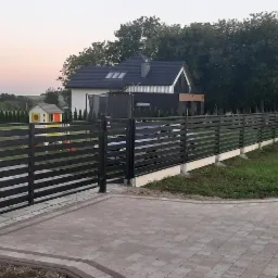 Nowoczesna, pozioma brama wjazdowa i ogrodzenie z metalowych profili w kolorze antracytowym, na tle domu z szarą dachówką i zieleni drzew, kostka brukowa na podjeździe.