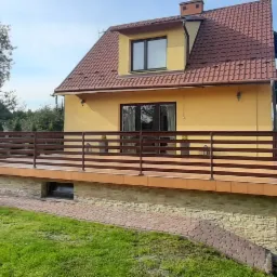 Żółty dom z brązowym dachem i balkonem z drewnianą barierką; widok z trawnika na elewację i otoczenie z zielenią.