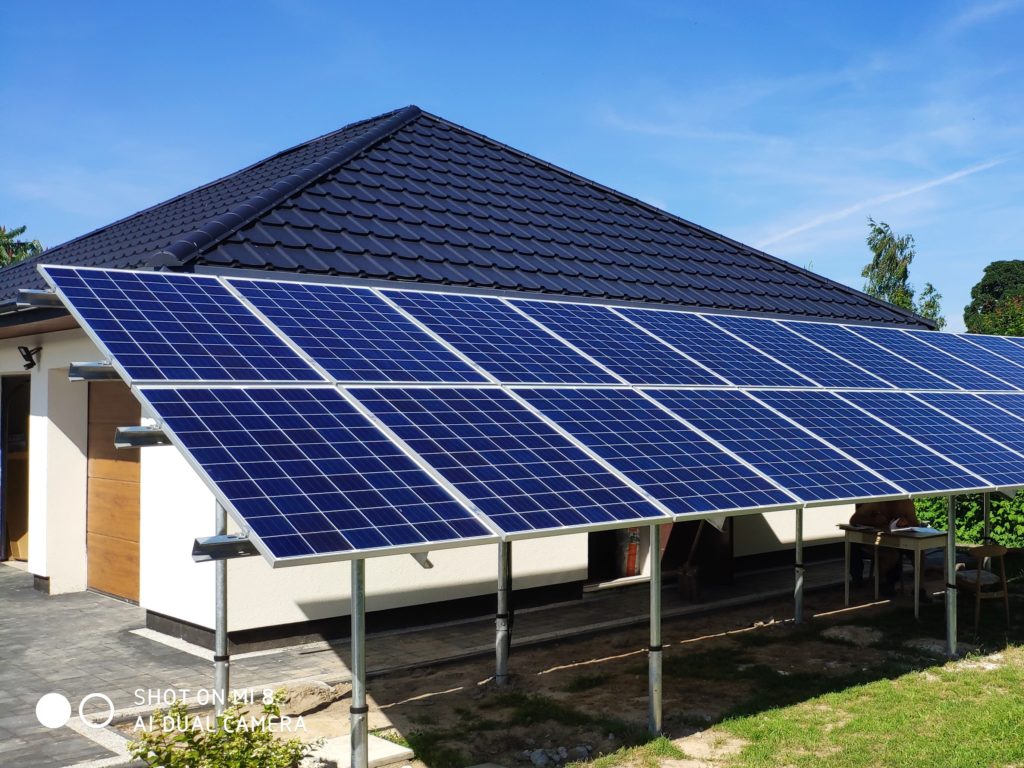 Panele słoneczne zamontowane na metalowych wspornikach, przylegające do budynku z ciemnym dachem i jasną elewacją, widoczne z poziomu trawnika w słoneczny dzień.