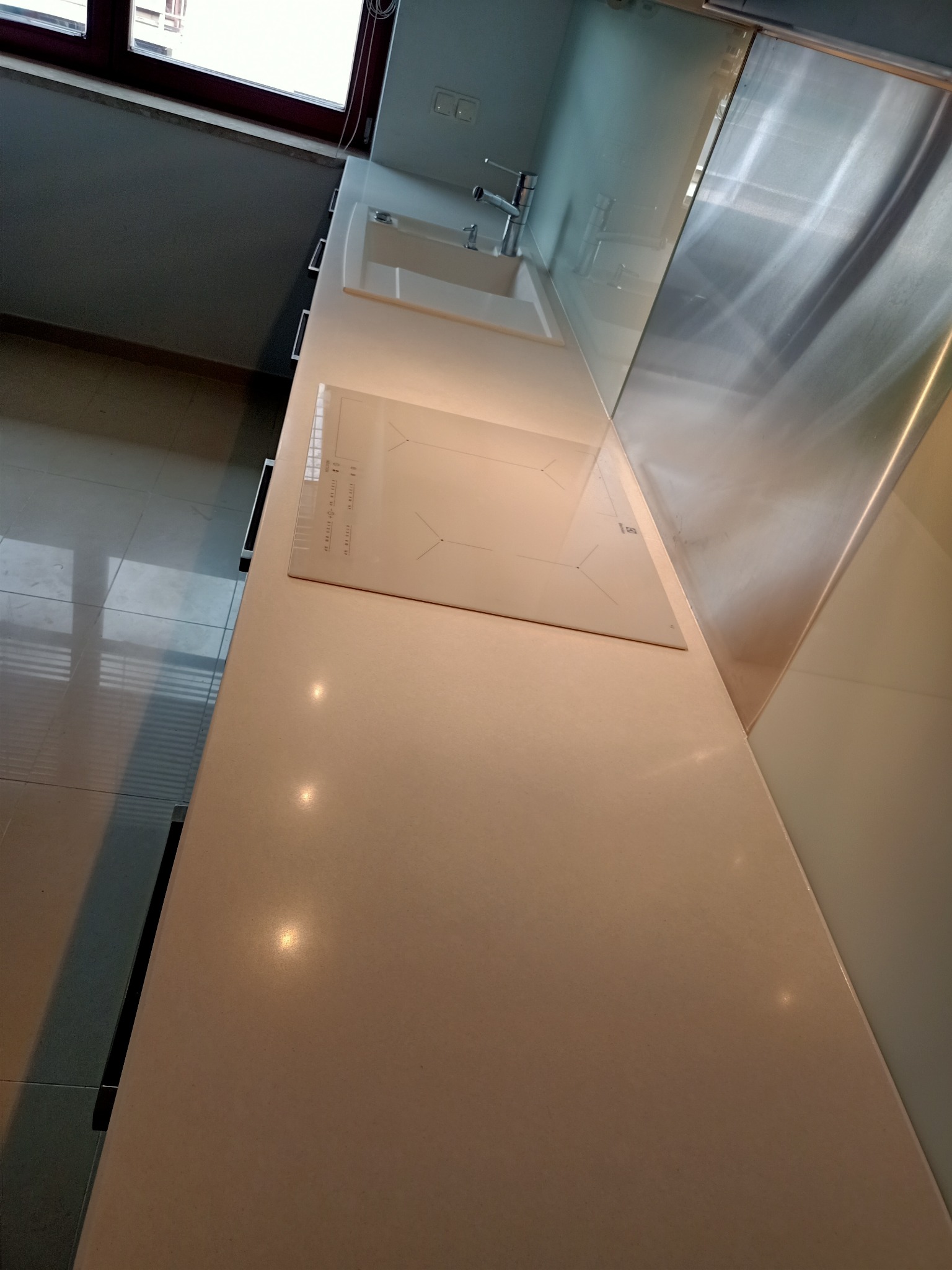 Jasny blat kamienny w kuchni z wbudowaną białą płytą indukcyjną i zlewozmywakiem, widok z góry, oświetlenie punktowe.