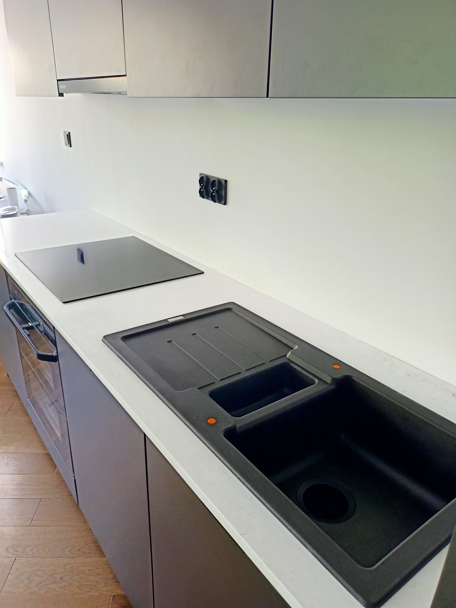 Nowoczesna kuchnia z białym blatem, czarnym zlewem kompozytowym z ociekaczem i płytą indukcyjną, minimalistyczne szafki w odcieniach szarości.