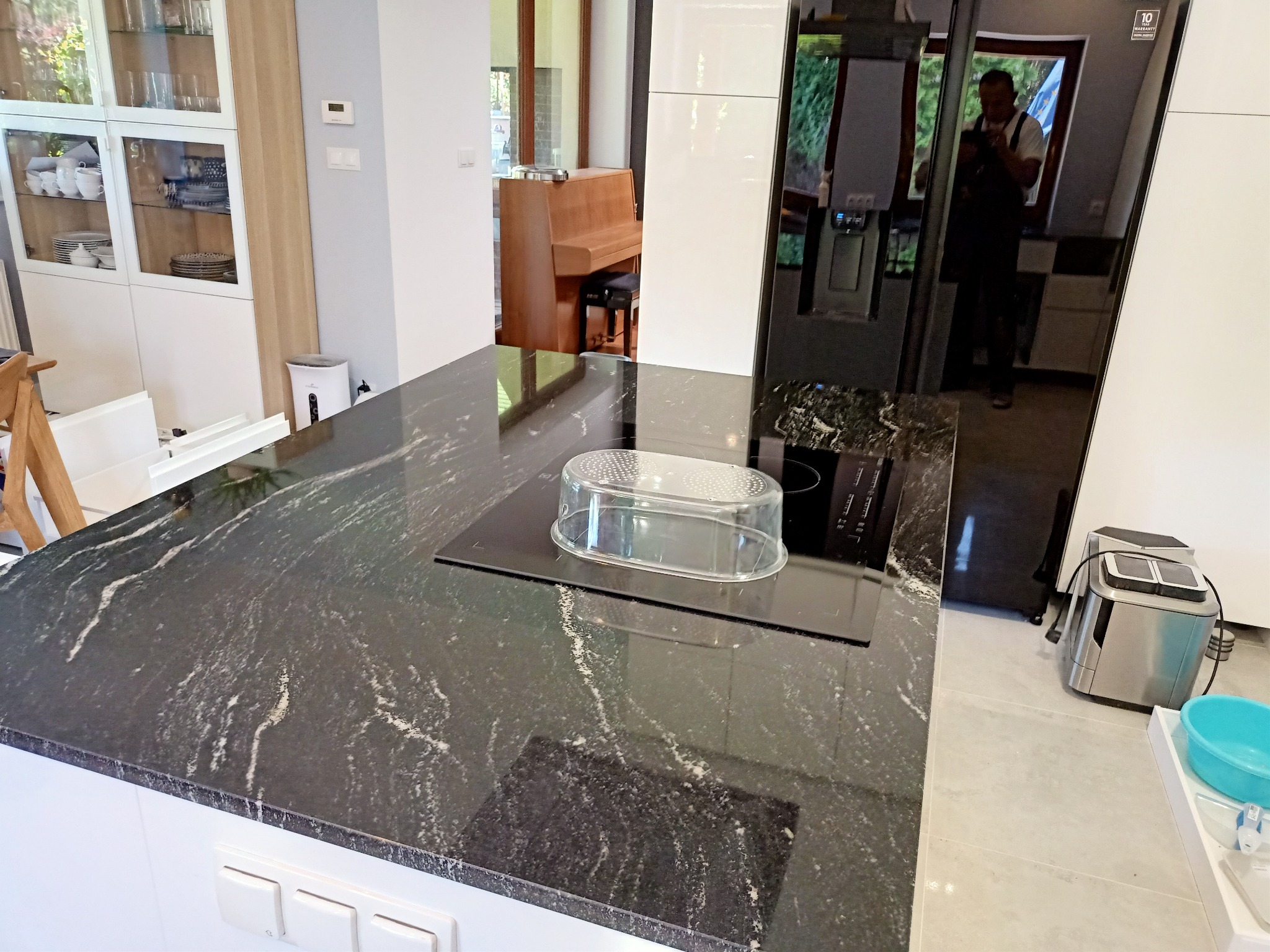 Czarny, błyszczący blat kuchenny z wyraźnym, białym użyleniem, z zamontowaną płytą indukcyjną przykrytą plastikową osłoną. W tle widoczna zabudowa kuchenna, pianino i odbicie osoby w lodówce.