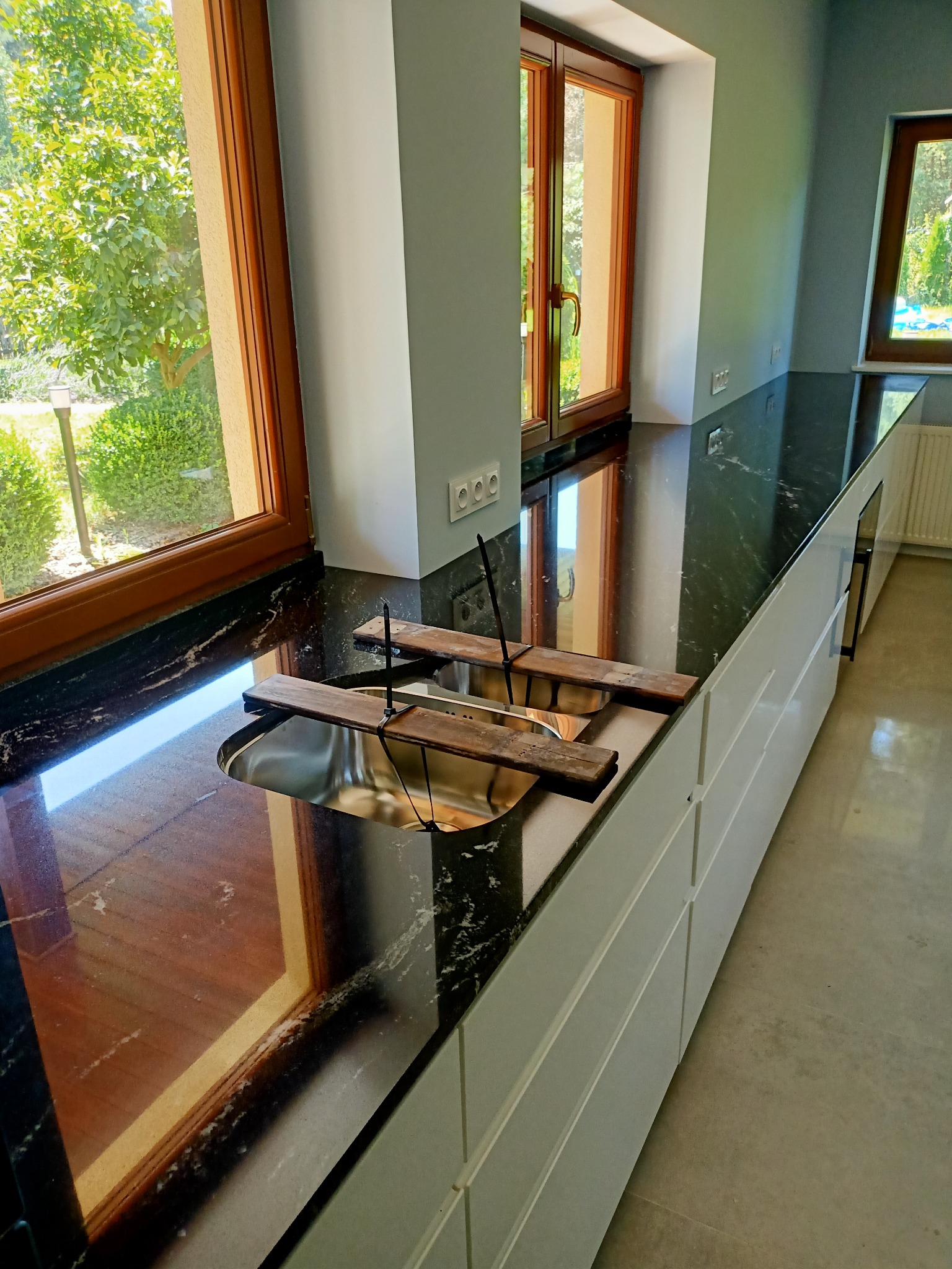 Czarny blat z kamienia naturalnego z wyciętym otworem na zlew, zabezpieczony drewnianymi listwami, wnętrze kuchni z oknami z widokiem na zieleń.