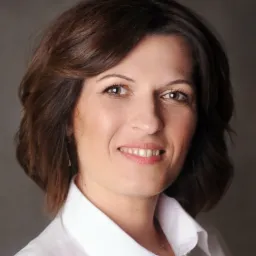 Portret uśmiechniętej kobiety z brązowymi włosami w białej koszuli na szarym tle.